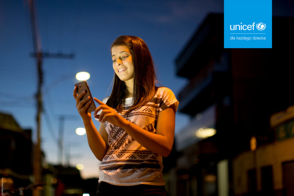 Dziewczynka ze smartfonem na tle miejskim, w górnym rogu logo INICEF Polska. Zdjęcie kampanii Przystań w sieci
