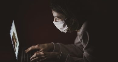 Kobieta w maseczce ochronnej siedząca w ciemnym pomieszczeniu, pisząca na laptopie.