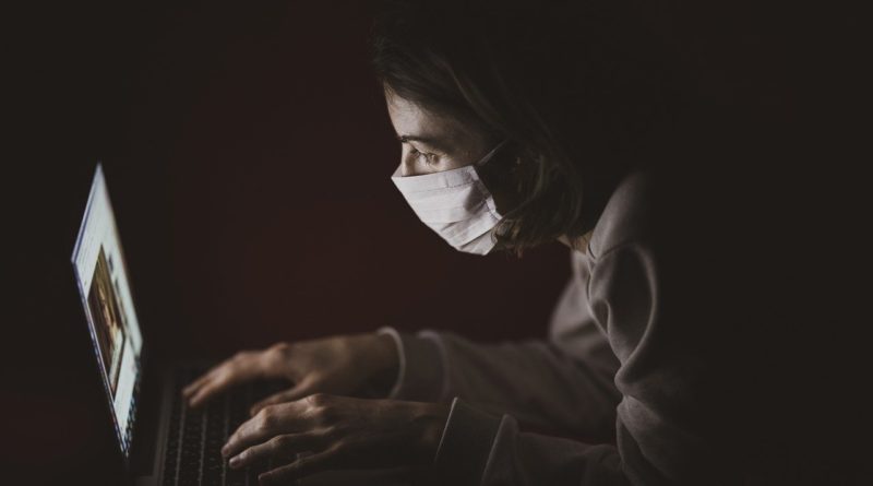 Kobieta w maseczce ochronnej siedząca w ciemnym pomieszczeniu, pisząca na laptopie.
