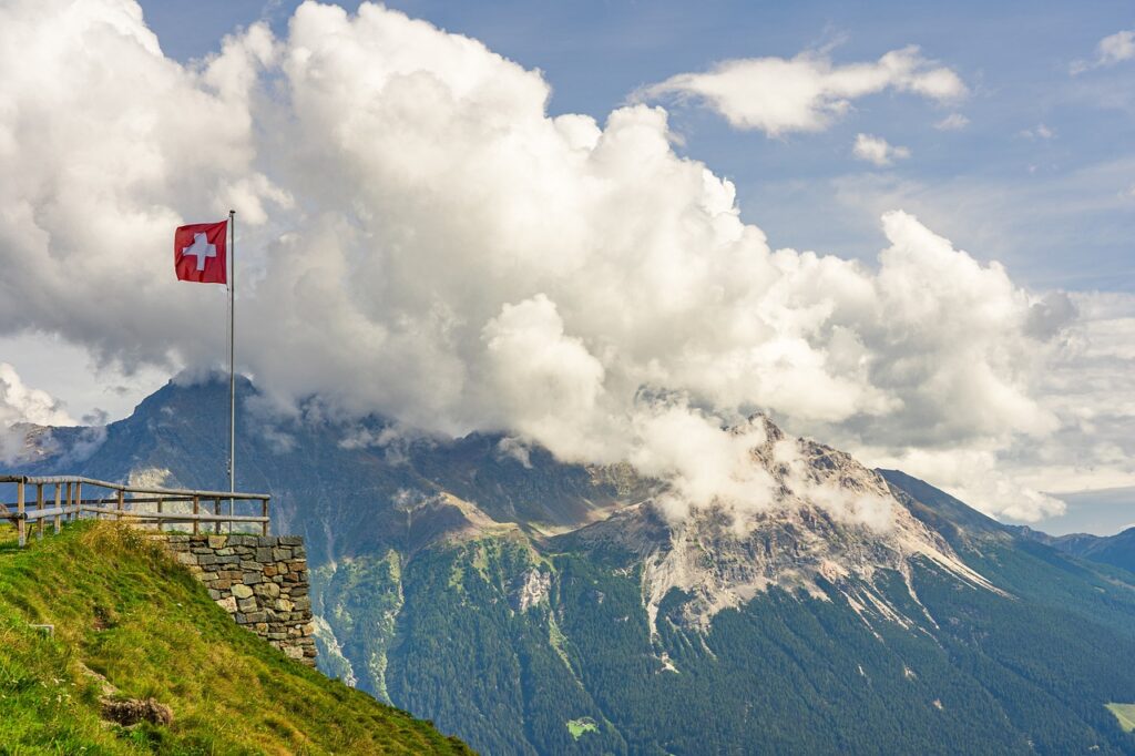 Widok na lekko ośnieżone, wysokie góry, okryte białymi chmurami na tle błękitnego nieba, po lewej stronie zdjęcia mała szwajcarska flaga, powiewająca na wietrze