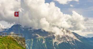 Widok na lekko ośnieżone, wysokie góry, okryte białymi chmurami na tle błękitnego nieba, po lewej stronie zdjęcia mała szwajcarska flaga, powiewająca na wietrze